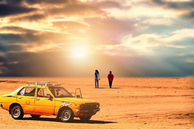 ludzie stojący na afrykańskiej pustynii, a obok stoi stary samochód o żółtym kolorze