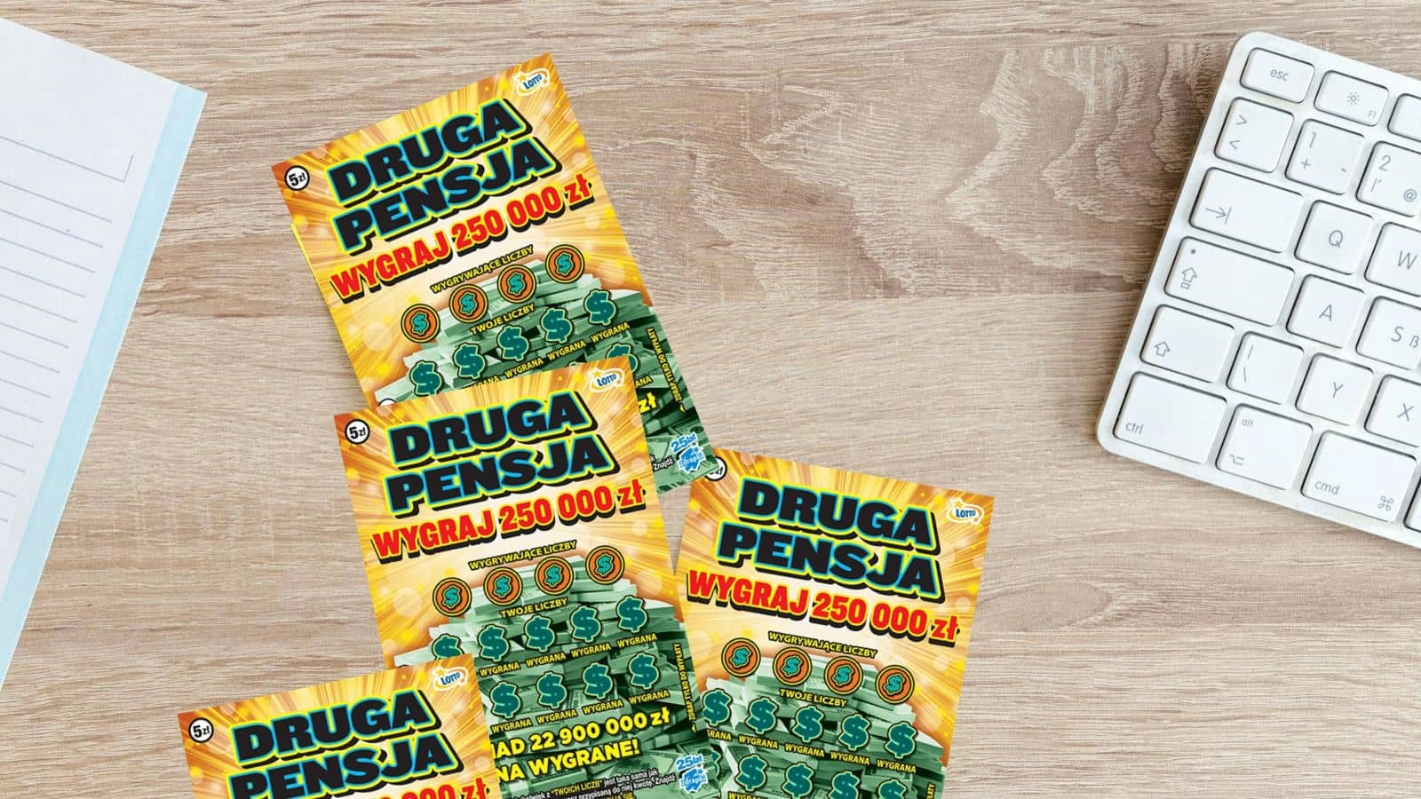 kilka zdrapek Lotto Druga Pensja, kartka z notesu, klawiatura na jasno drewnianym blacie stołu
