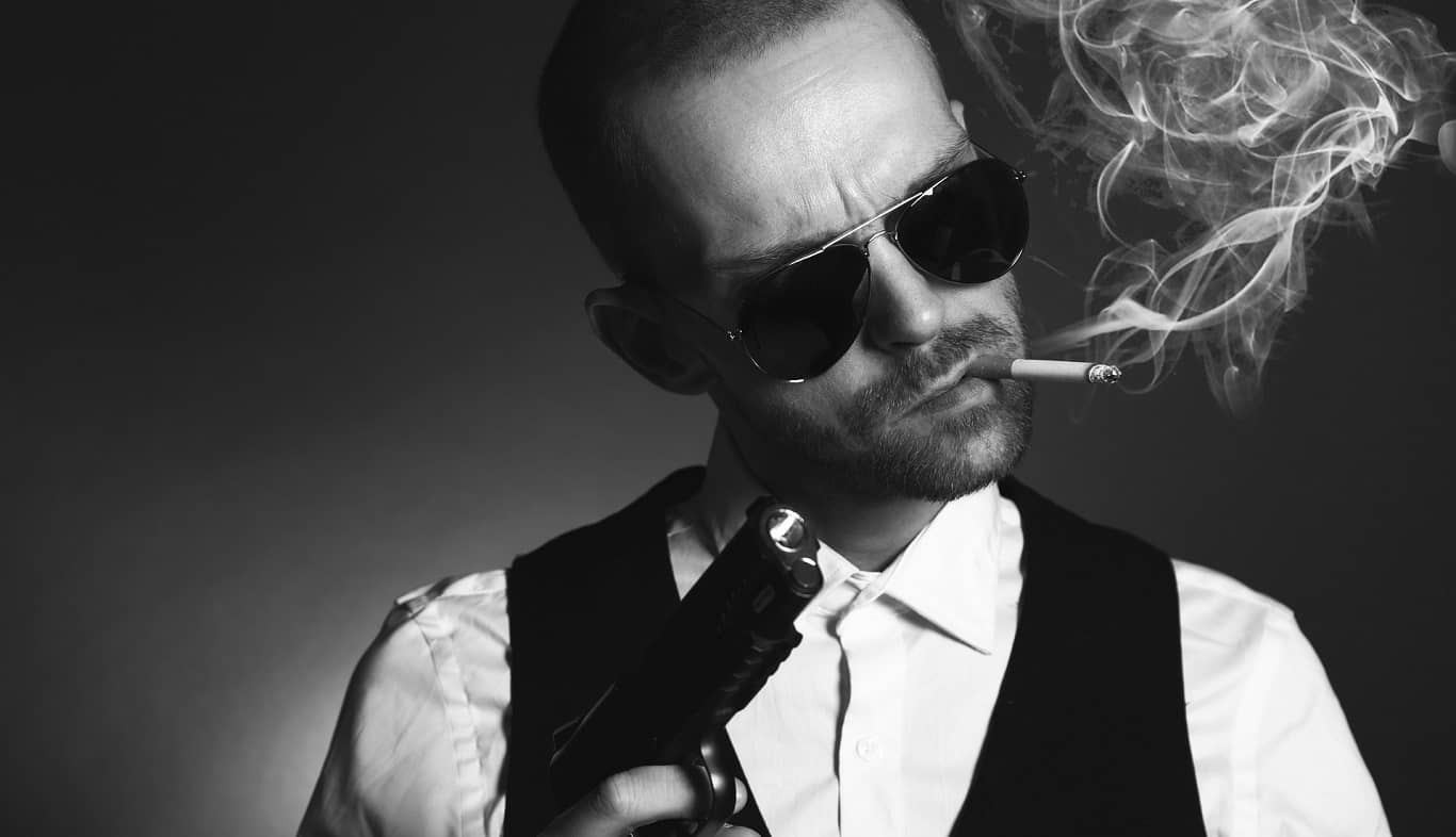 mężczyzna w białej koszuli, okularach przeciwsłonecznych i kamizelce trzyma pistolet i pali papierosa

