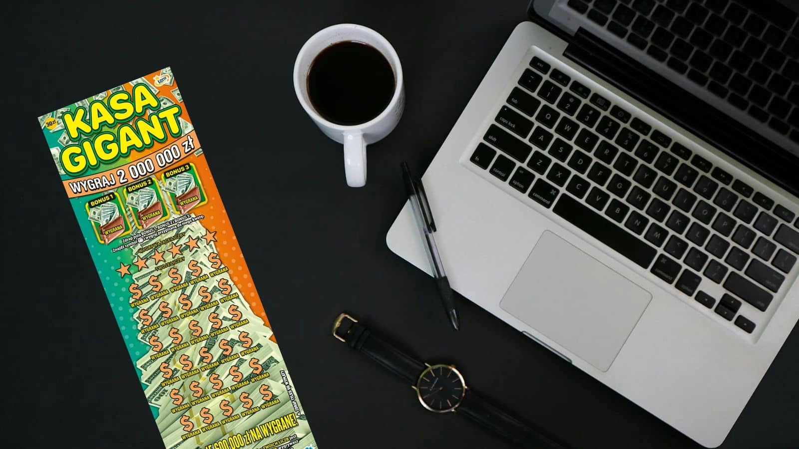 zdrapka lotto Kasa Gigant obok zegarka, kubka z kawą i macbooka na czarnym blacie biurka, widok z góry