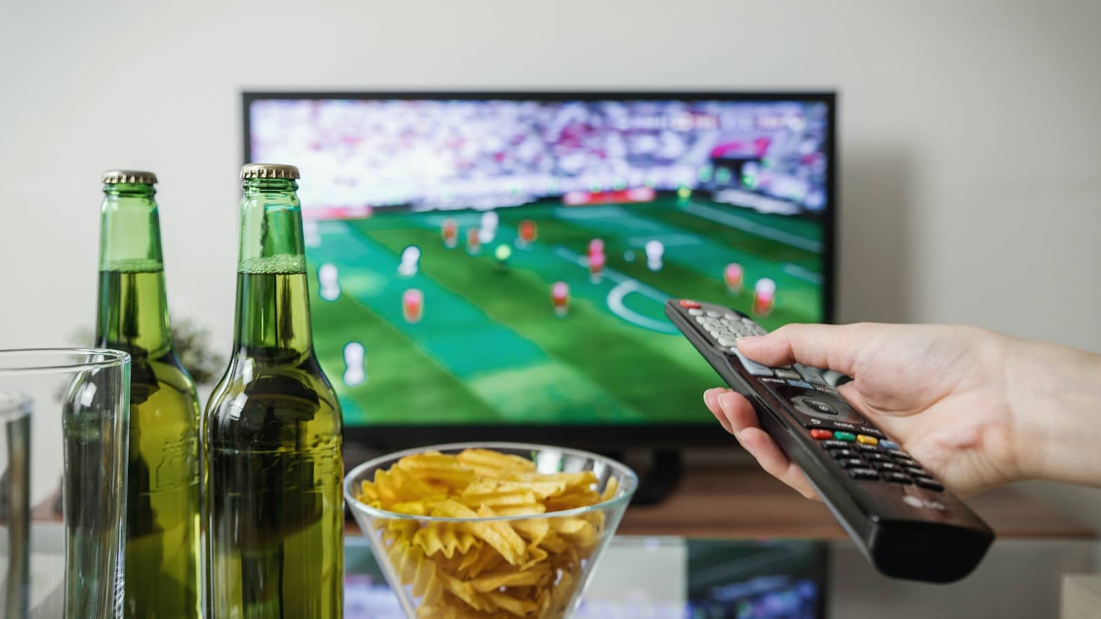dłoń trzymająca pilot od tv, obok stoi miska z chipsami i dwa piwa, w tle duży telewizor transmitujący mecz piłki nożnej