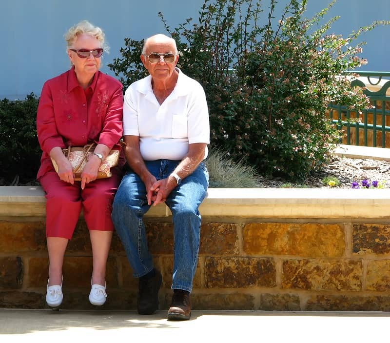 starsza kobieta ubrana na czerwono i starszy pan ubrany na biało-niebiesko siedzą na murku oświetleni słońcem 