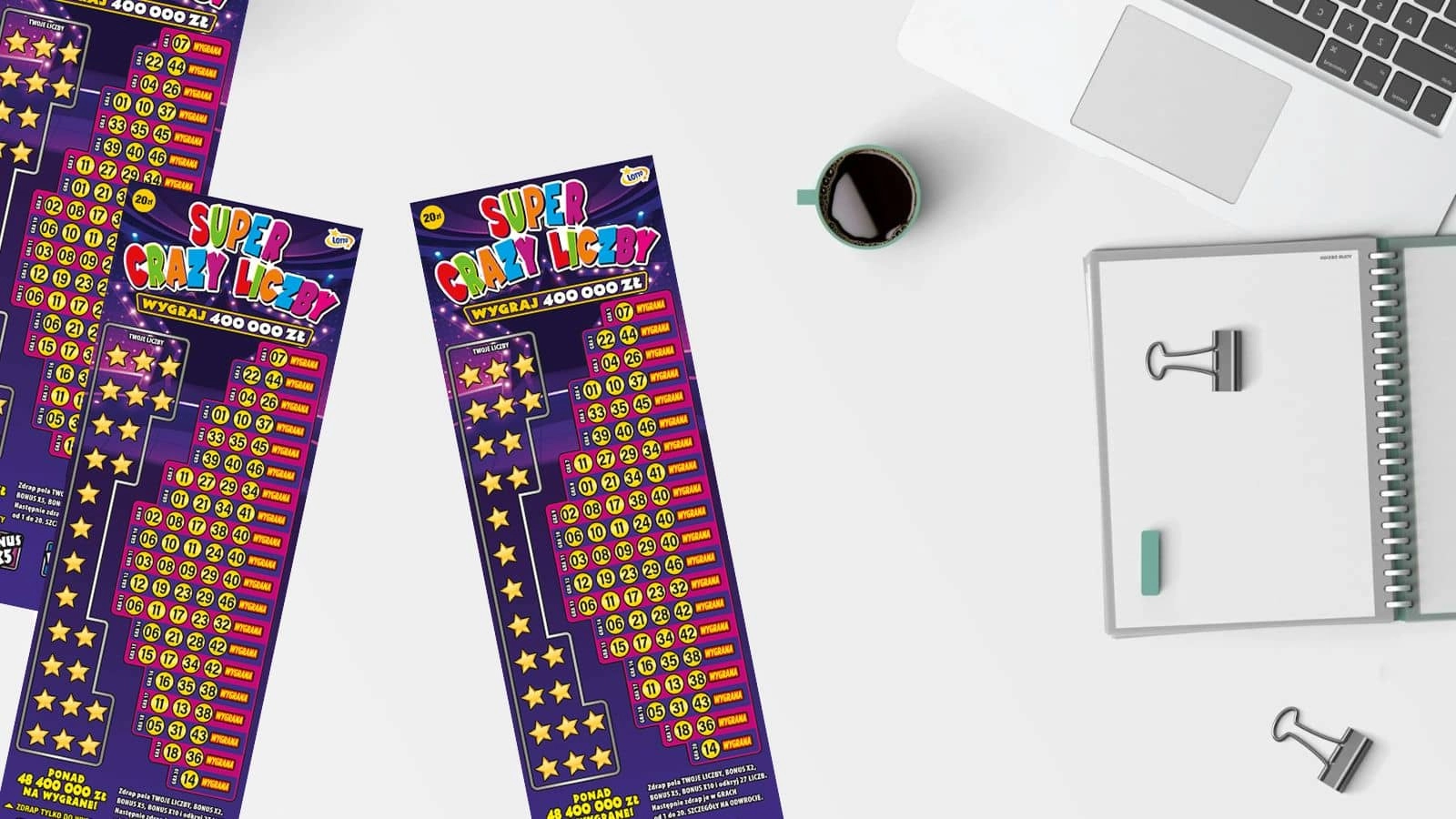 zdrapki Lotto Super Crazy Liczby oraz filiżanka kawy, zeszyt, klamry biurowe, laptop na białym blacie stołu, widok z góry